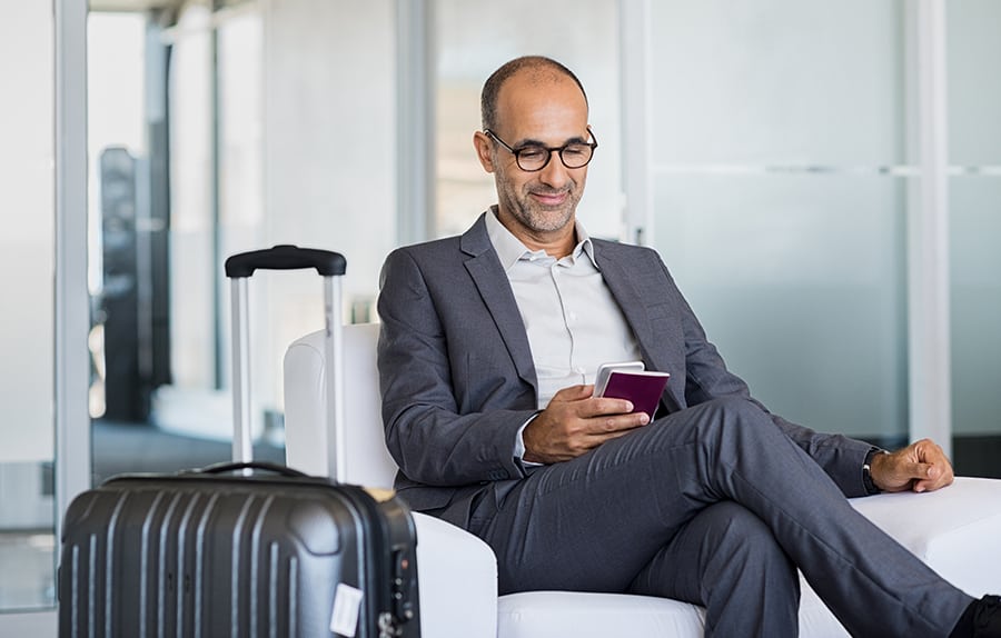 Mature businessman using mobile phone at the airport in the waiting room. Business man typing on smartphone in lounge area. Portrait of latin man sitting and holding passport with luggage.