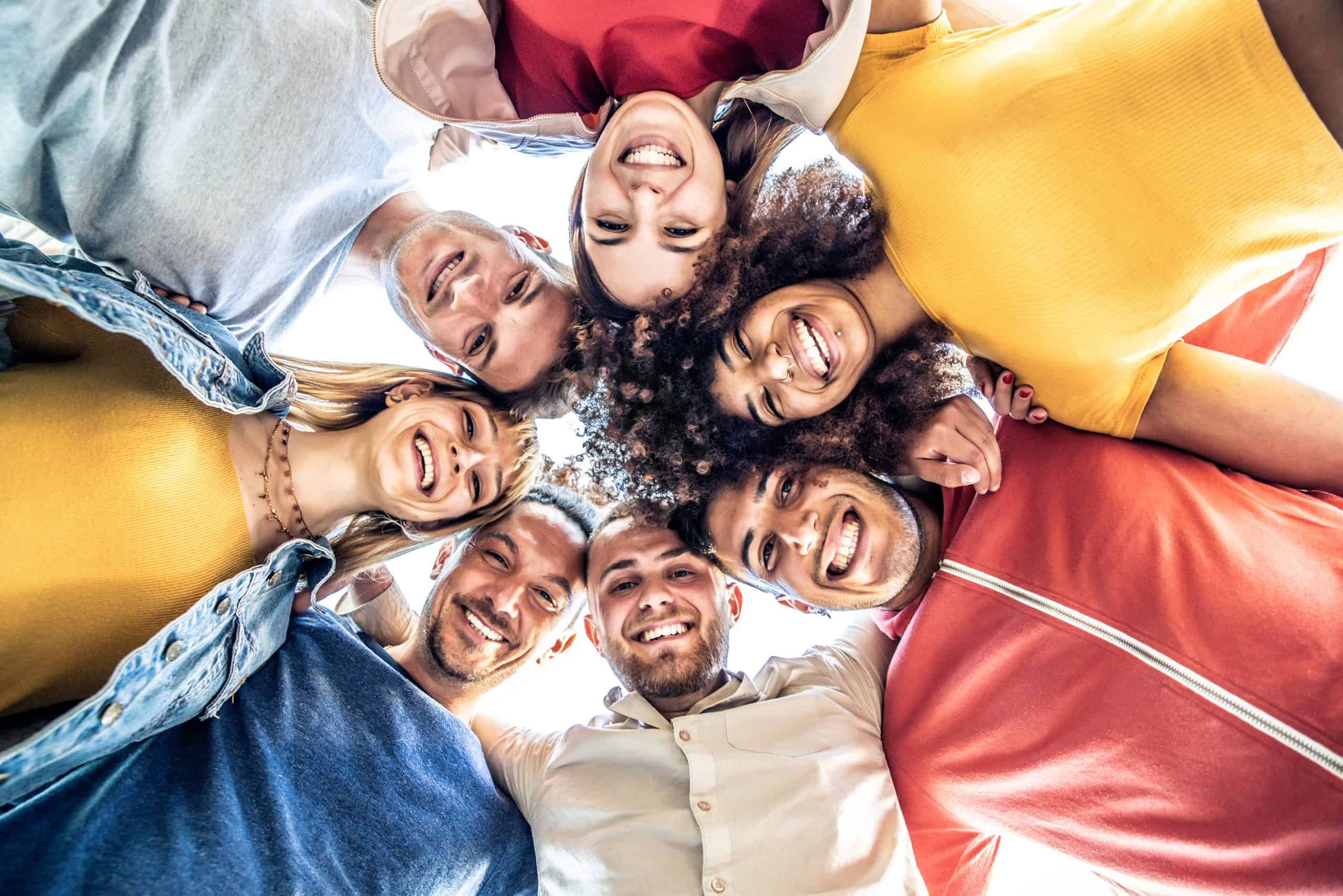 Multiracial group of young people standing in circle and smiling at camera - Happy diverse friends having fun hugging together - Low angle view